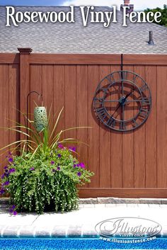 a clock mounted to the side of a wooden fence next to a planter filled with flowers