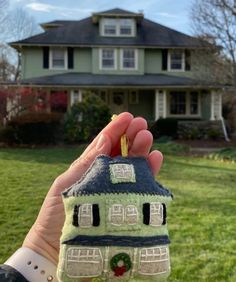 a hand holding up a small house ornament in front of a green house