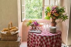 a table with food and wine glasses on it in front of a window, next to two wicker baskets filled with flowers