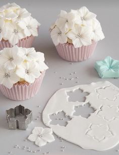 cupcakes decorated with white flowers and icing are on a table next to cookie cutters