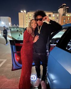 a man and woman standing in front of a car at night with their arms around each other