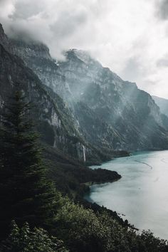 a lake surrounded by mountains and trees under a cloudy sky with water in the foreground