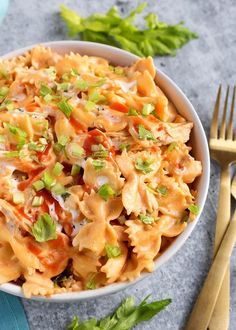 a white bowl filled with pasta and vegetables next to gold forks on a gray surface