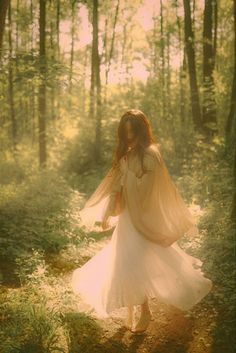 a woman in a white dress walking through the woods with her hair blowing in the wind