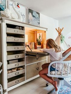 a woman sitting at a desk in front of a cabinet with many drawers and shelves