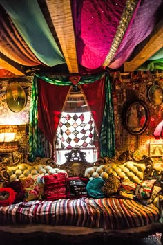 a bed covered in lots of pillows and blankets next to a window with colorful drapes