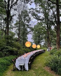 a long table is set up in the woods for an outdoor dinner party with paper lanterns hanging from the trees