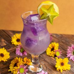 a glass filled with purple liquid surrounded by yellow and pink flowers on a wooden table