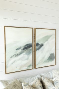 two paintings hanging on the wall above a couch in a room with white walls and pillows