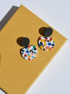 a pair of colorful earrings sitting on top of a yellow book next to a black button