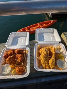 two takeout containers filled with fried chicken and french fries next to a boat on the water