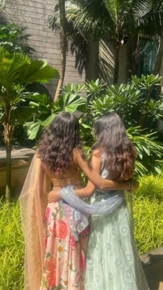 two women in dresses standing next to each other near some trees and bushes with palm trees behind them