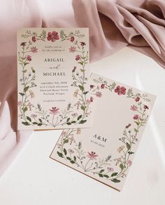 two wedding cards with flowers on them sitting next to a pink satin material draped in the background