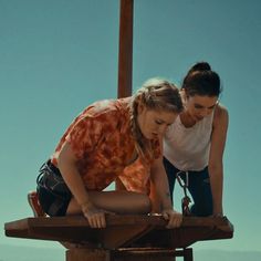 two women are looking at something on a table outside in the day time and one is leaning over another woman's shoulder