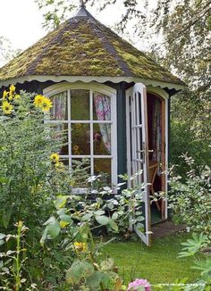 a small garden shed with a green roof and white windows in the middle of some flowers