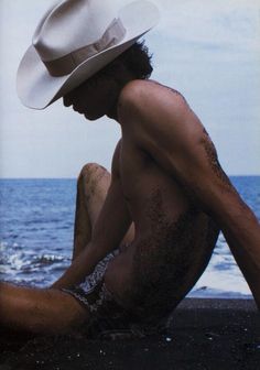 a shirtless man sitting on the beach with his hat over his head and body covered in sand