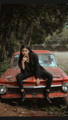 a woman sitting on top of an old red car in the woods with her hand under her chin