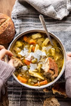 two hands holding a bowl of soup with bread on the side