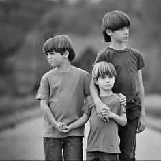 three young boys standing next to each other on a road with trees in the background