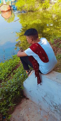 a young boy sitting on the edge of a lake looking at something in the water