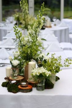 an arrangement of flowers and candles on a long table with white clothed tables in the background