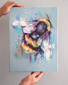 two hands holding up a painting of a bee on a blue background with multicolored spots