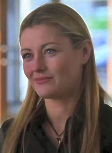 a woman with long blonde hair wearing a black shirt and looking at the camera while standing in front of a window