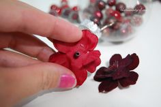 a hand holding a red teddy bear next to a flower and glass vase filled with beads