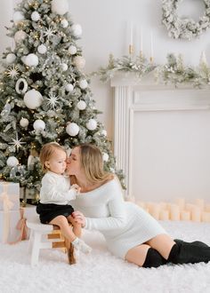 a mother kissing her baby while sitting in front of a christmas tree with presents on the floor