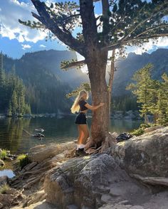 a woman standing next to a tree on top of a rocky hillside near a lake