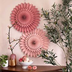 two pink paper flowers are hanging on the wall next to a bowl of fruit and greenery