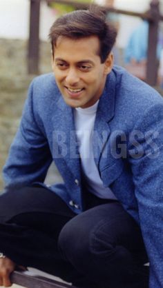 a man in a blue jacket and black pants sitting on a bench smiling at the camera