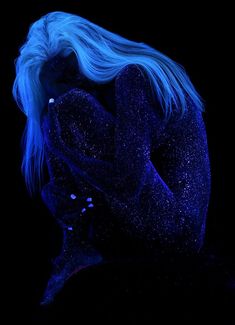 a woman with blue hair is sitting in the dark and has her hands on her face