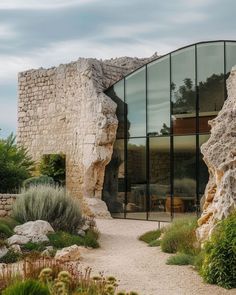 a stone building with glass walls and plants in the foreground, next to a gravel path