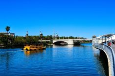 a yellow boat traveling down a river next to a bridge and palm trees on the other side