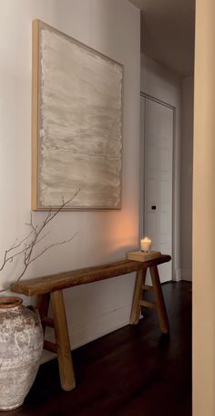 a painting hangs on the wall next to a wooden bench and vase with a candle in it