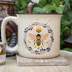 a mug with a bee painted on it sitting next to some honey jars and plants