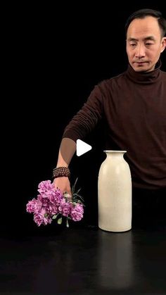 a man holding flowers in front of a white vase on a black table with purple flowers