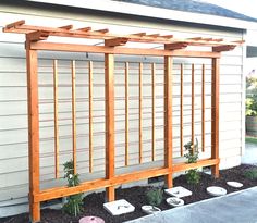 a wooden trellis next to a house with rocks and plants in the foreground