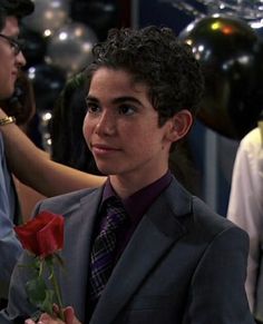 a young man in a suit and tie holding a rose with other people around him