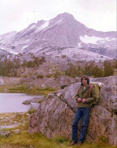 a man standing on top of a large rock next to a body of water with mountains in the background