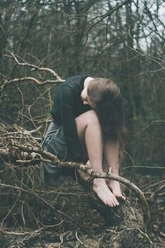 a woman sitting on top of a tree branch in the middle of a forest with her head down