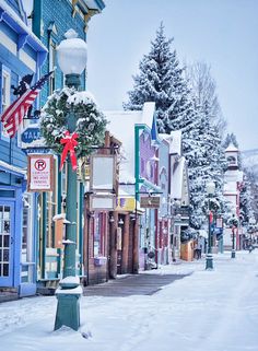 the street is covered in snow and has many shops on each side with christmas decorations