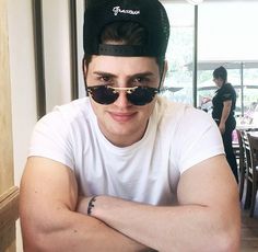 a young man wearing sunglasses and a hat sitting at a table with his arms crossed