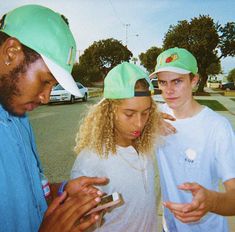 three young people standing in the street looking at their cell phones while one looks at his phone