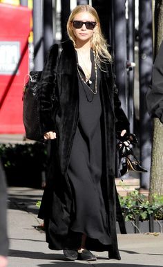 a woman walking down the street in a long black coat and dress with sunglasses on her head