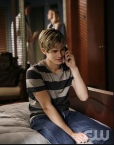 a young man sitting on top of a bed talking on a cell phone while another person stands in the background