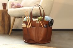 a brown leather purse with knitting needles and yarn in it sitting on the floor next to a chair