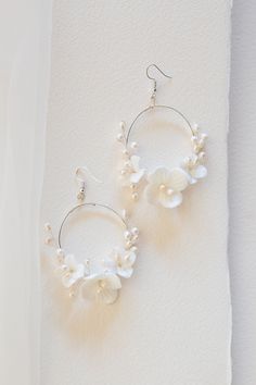 The Flora Hoops have a modern look with the aura of an heirloom. These romantic circular hoop earrings are feminine, yet bold. Decorated with light ivory ceramic blossoms and natural freshwater pearls, the Flora Earrings would be a stunning compliment to any bridal look. Details: Available finishes: gold/ Light Ivory or silver/ light ivory Ceramic Blossom color: light ivory Gold/ Silver Plated jewelry wire Attached to gold/ silver plated ear wire hooks Dimensions: 2.25x3 inches Final Sale White Flower-shaped Earrings With Pearl Charm, Delicate White Hoop Earrings With Pearl Charm, White Small Hoop Pearl Earrings, Elegant White Flower Hoop Earrings, White Small Hoop Pearl Earrings With Ear Wire, White Hoop Earrings With Pearl Charm For Wedding, White Pearl Charm Hoop Earrings For Wedding, White Pearl Small Hoop Earrings With Ear Wire, Elegant Adjustable White Hoop Earrings