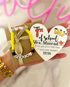 a hand holding a heart shaped card with two bracelets attached to it and a tag that says happy 1st day of school